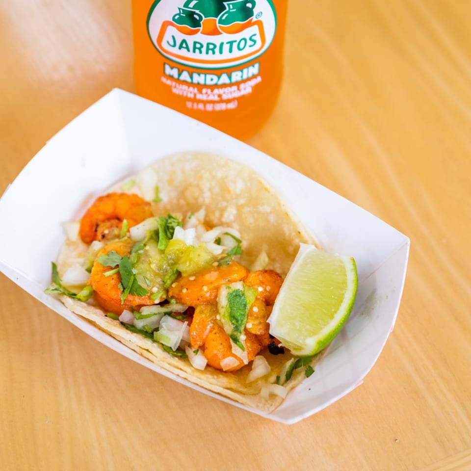 Shrimp taco with salsa verde, onions, cilantro, and a lime wedge, next to a Jarritos soda.