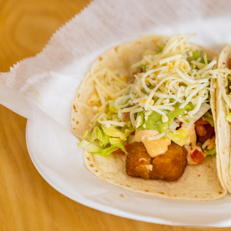 Two Blue Tortilla fish tacos with shredded lettuce, cheese, and sauce on a white plate.