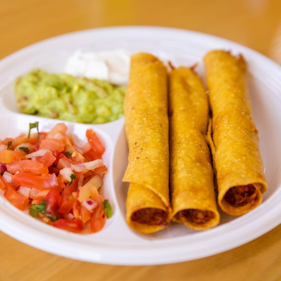 Three golden taquitos with guacamole, sour cream, and pico de gallo on a white plate.
