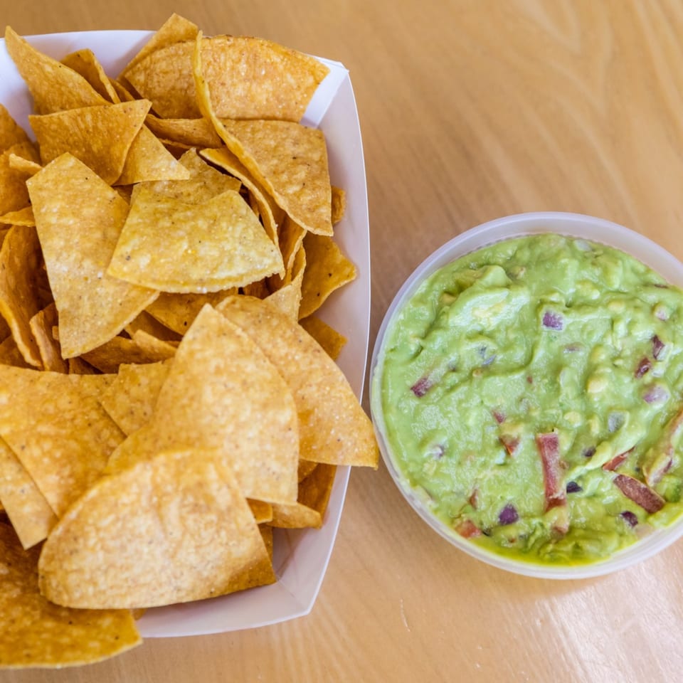 A white container filled with tortilla chips next to a bowl of guacamole on a wooden table.